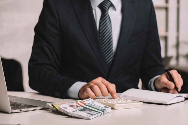 cropped shot of businessman using calculator and taking notes in notepad at workplace with laptop and russian rubles banknotes  