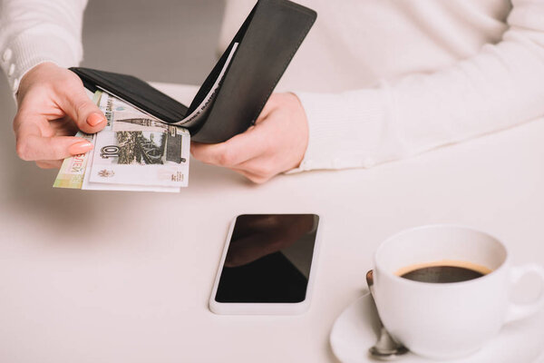 cropped shot of woman holding wallet and russian rubles banknotes above white table with cup of coffee and smartphone