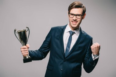 handsome businessman in glasses celebrating victory and holding trophy in hand on grey background clipart