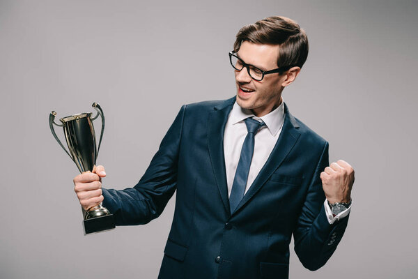 cheerful businessman in glasses celebrating victory and holding trophy in hand on grey background