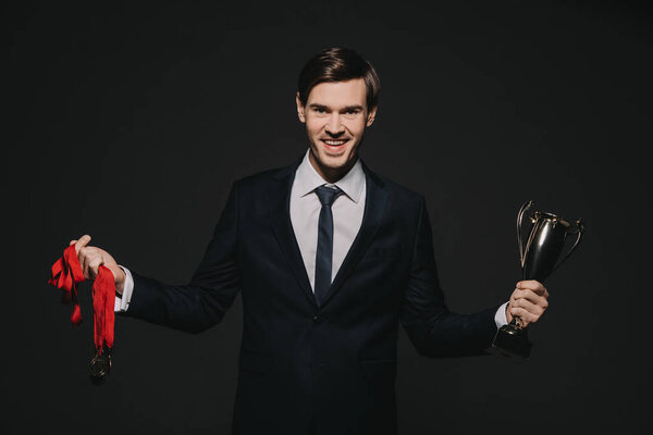 smiling businessman holding medals trophy in hands isolated on black