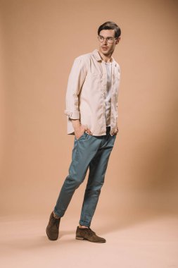 elleri ceplerinde bej renkli arka plan üzerinde durup gözlüklü kendine güvenen adam