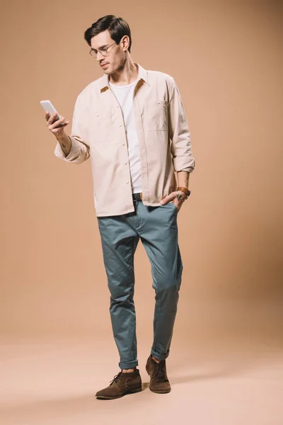 Stilig Man Glasögon Tittar Smartphone Stående Beige Bakgrund — Stockfoto