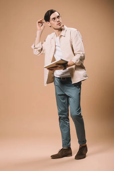 英俊的男人在眼镜举行笔记本和笔 而站在米色的背景 — 图库照片