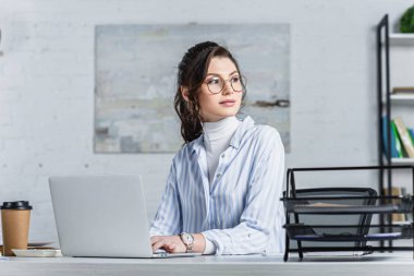 Dizüstü bilgisayar kullanarak ve uzağa bakarak bardaklarda çekici iş kadını 