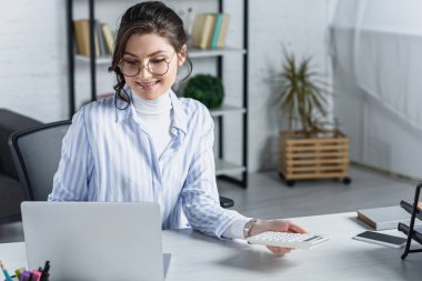 attractive woman in glasses holding calculator near laptop in modern office  clipart