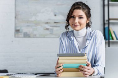 cheerful woman sitting near books in modern office  clipart