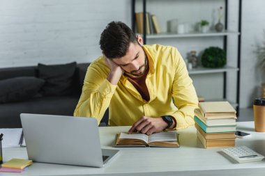 tired man studying with book near laptop in modern office  clipart
