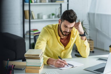 focused man writing in notebook near laptop and books in modern office  clipart