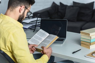 bearded man studying with book near laptop with blank screen in modern office clipart