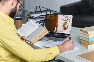 bearded man studying with book near laptop with tickets online website on screen in modern office clipart