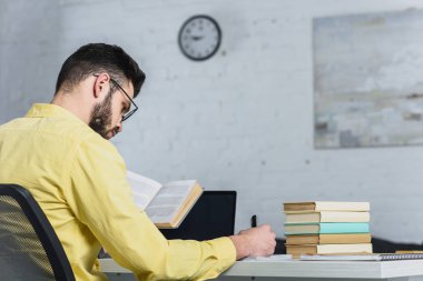 selective focus of man writing while holding book in modern office clipart