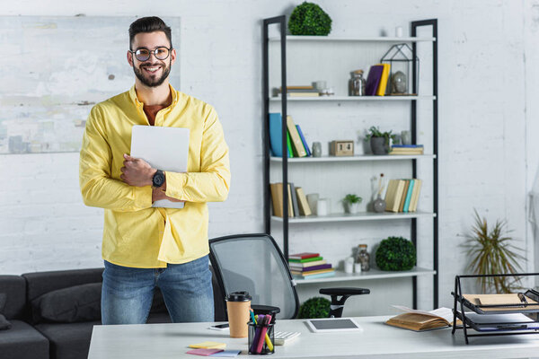 Бородатый бизнесмен в очках держит цифровой планшет и смотрит в камеру на рабочем месте
