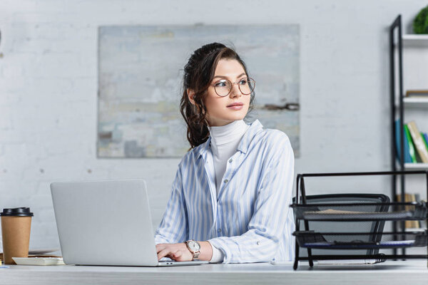 Attractive businesswoman in glasses using laptop and looking away 