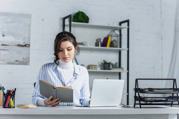 attractive woman studying with book near laptop in modern office 