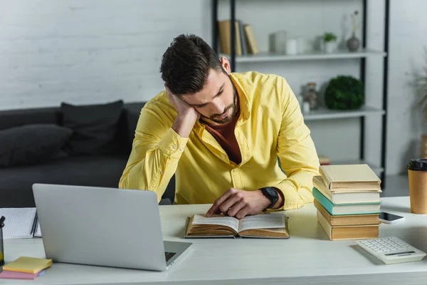 tired man studying with book near laptop in modern office