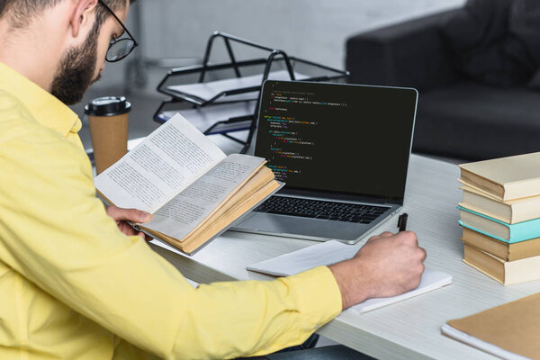 bearded man studying with book near laptop with computer coding on screen in modern office