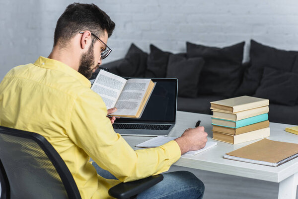 bearded man studying with book near laptop with blank screen in modern office