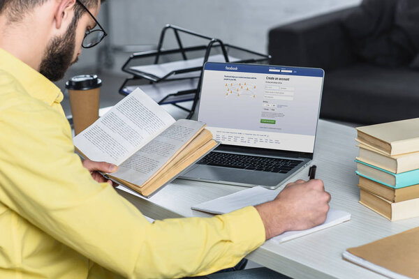 bearded man studying with book near laptop with facebook website on screen in modern office