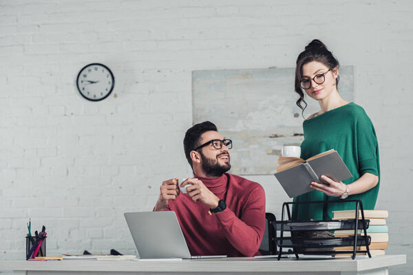cheerful man in glasses looking at woman studying with book and holding cup with drink