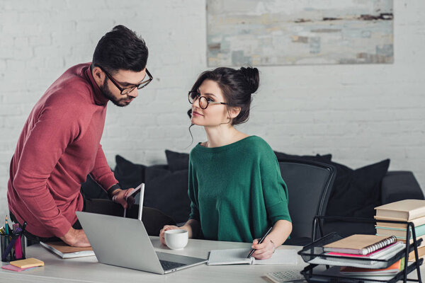 attractive woman in glasses writing in notebook and looking at coworker 