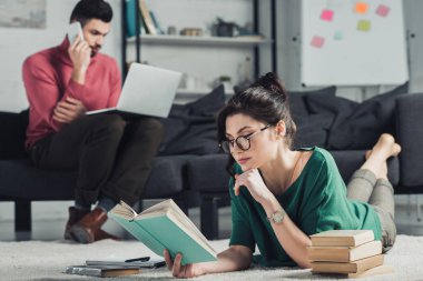 seçici odak halı üzerinde yalan ve arka plan üzerinde dizüstü bilgisayar kullanan yakışıklı adam yakınındaki kitapla okuyan kadın