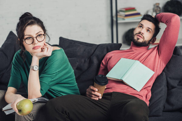 bored woman in glasses holding apple and sitting near handsome man with paper cup