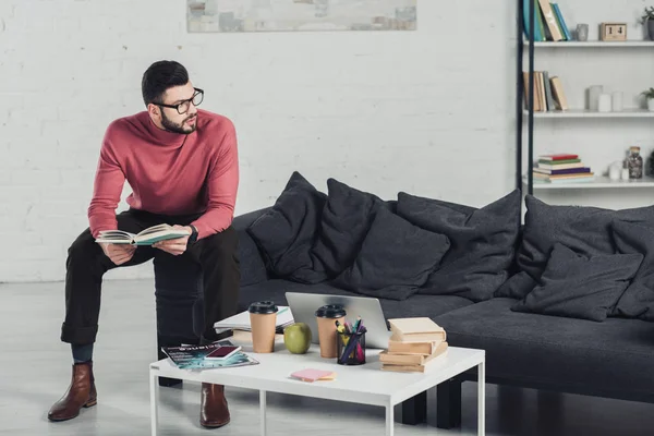 Hombre Guapo Sosteniendo Libro Sentado Sofá Cerca Mesa Café — Foto de Stock