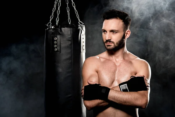 thoughtful man standing with crossed arms near punching bag on black with smoke