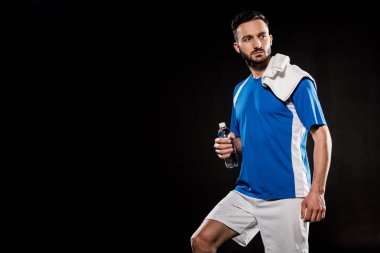 tek tip havlu omuz ile ayakta ve siyahta izole su ile şişe tutan yakışıklı adam