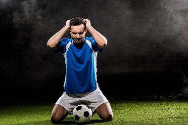 handsome football player holding head while sitting on grass on black with smoke clipart
