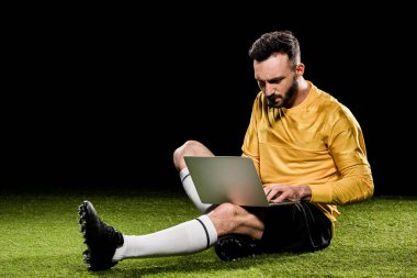 handsome referee holding whistle in mouth and looking at laptop while sitting on grass isolated on black clipart
