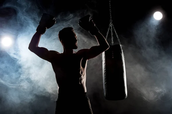 stock image silhouette of muscular athlete in boxing gloves with hands above head on black with smoke