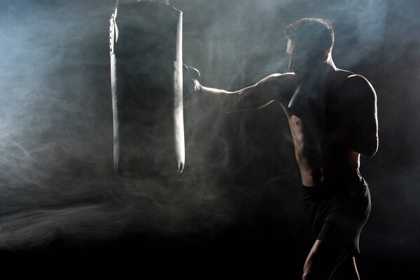 silhouette of sportsman in boxing gloves hitting punching bag on black with smoke
