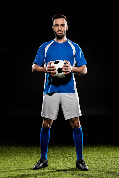 футболист, стоящий с мячом на зеленой траве, изолированный на черном
