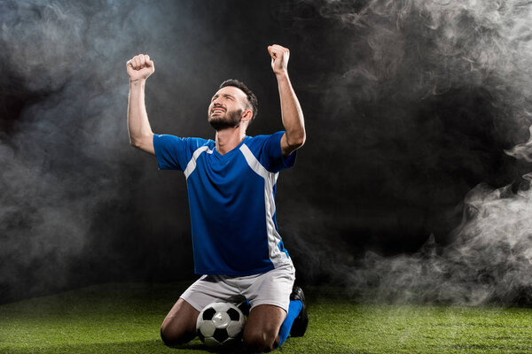 красивый футболист празднует победу, сидя на траве на черном от дыма
