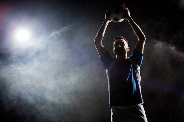 silhouette of cheerful football player holding ball above head on black with smoke  