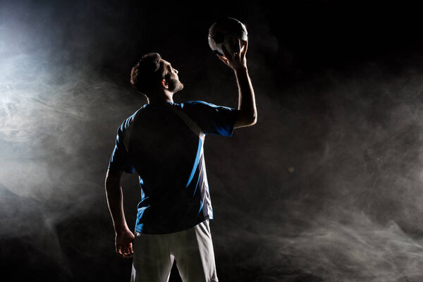 silhouette of player in uniform holding ball above head on black with smoke  