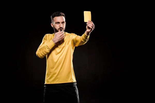 handsome referee showing yellow card and blowing whistle isolated on black
