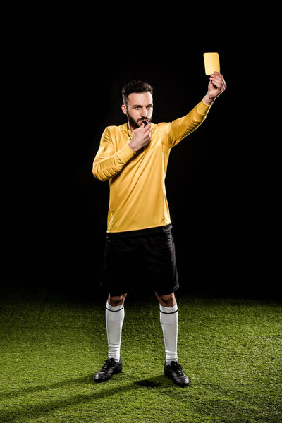 handsome referee standing on grass and showing yellow card while blowing whistle isolated on black
