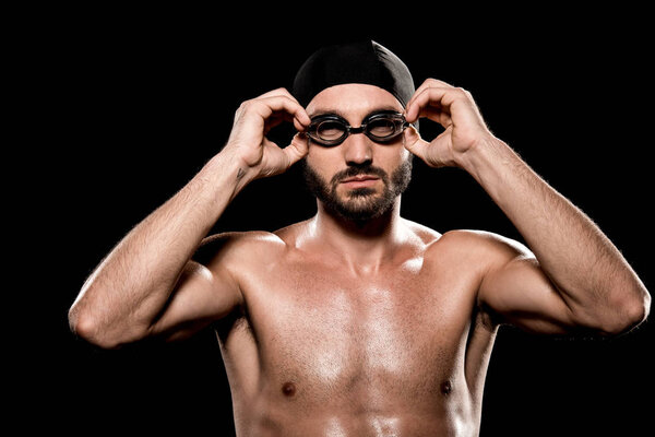 confident swimmer standing in swimming cap and touching goggles isolated on black 