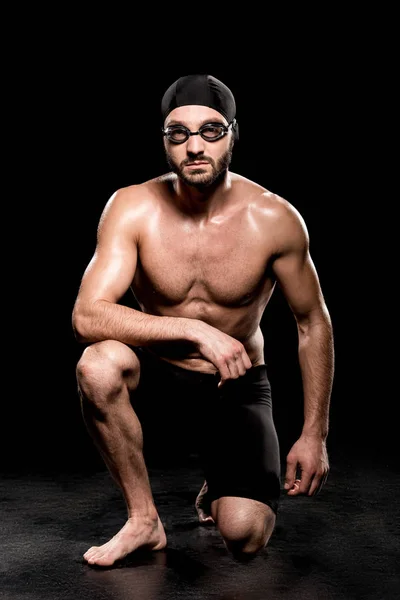 athletic swimmer sitting in swimming cap and goggles on black background