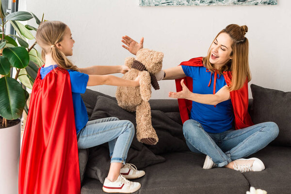 Attractive mother and cute daughter in red cloaks playing with teddy bear at home
