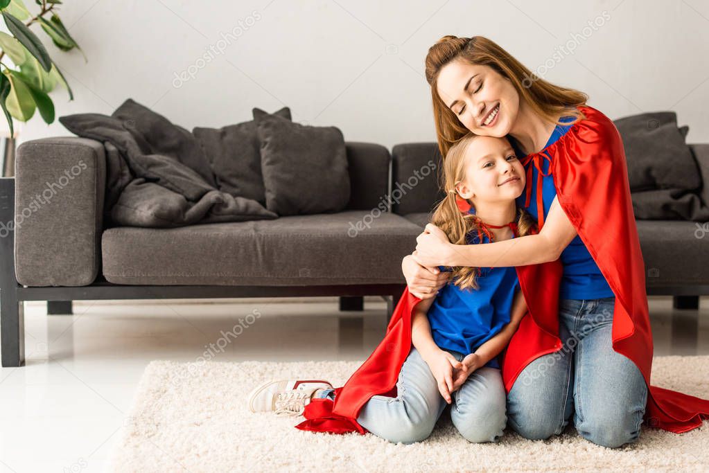 Kid and mother in red cloaks sitting on floor and hugging at home