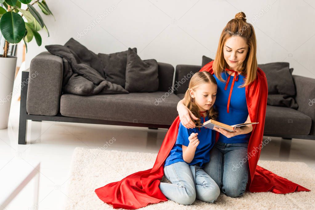Cute kid and mother in red cloaks sitting on floor and reading book