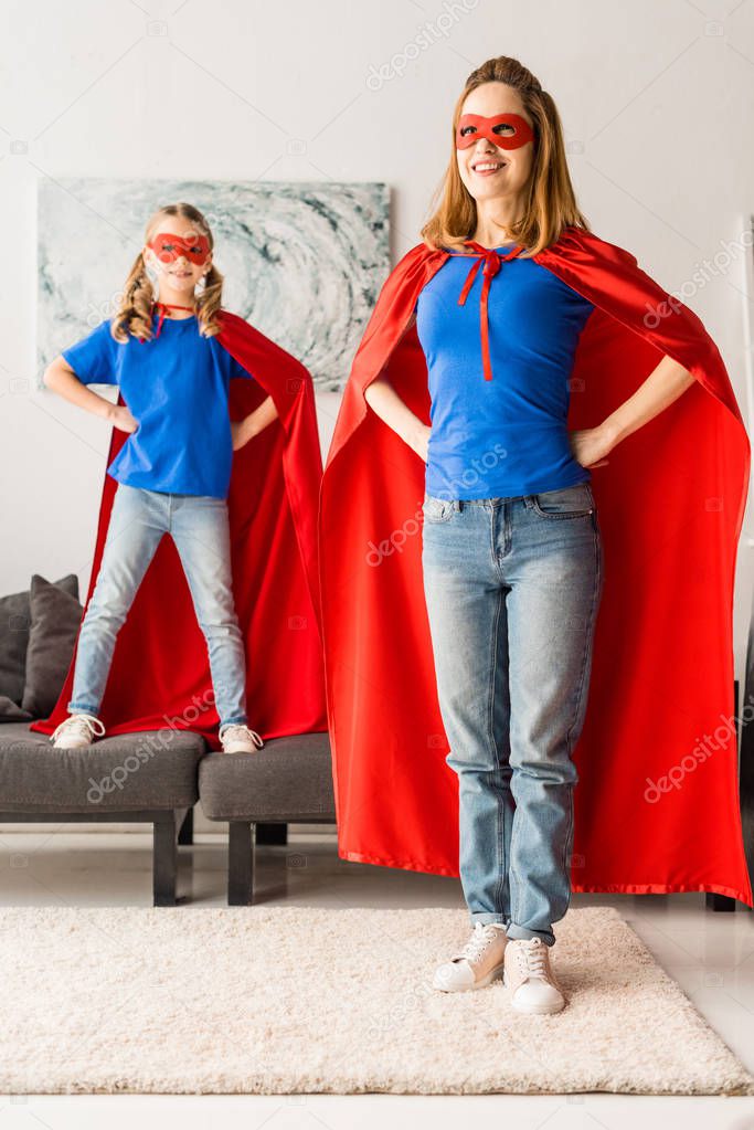 Cute daughter and mother in red cloaks and masks standing with hands on hips