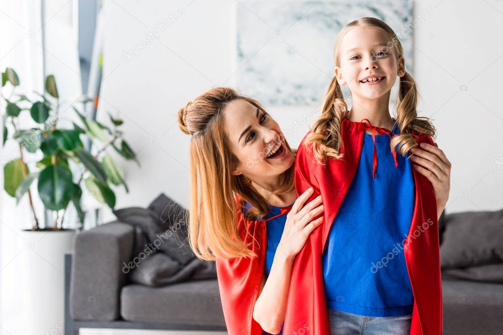 Cute kid and attractive mother in red cloaks smiling and hugging at home