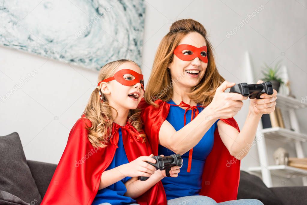 Smiling mother and daughter in red masks and cloaks playing video game