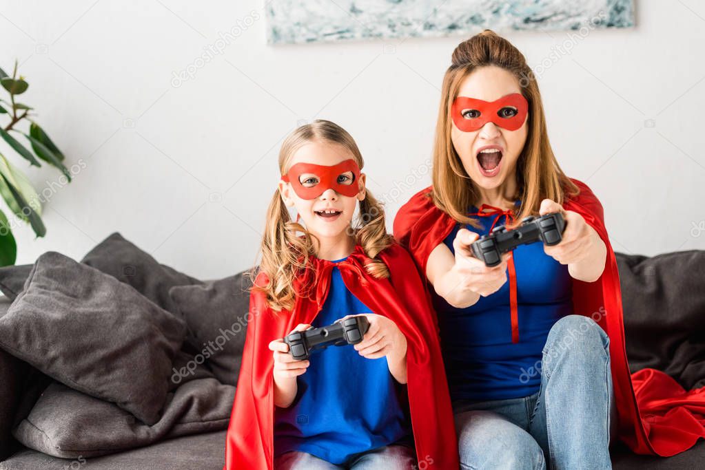 Beautiful mother and daughter in red masks and cloaks playing video game at home