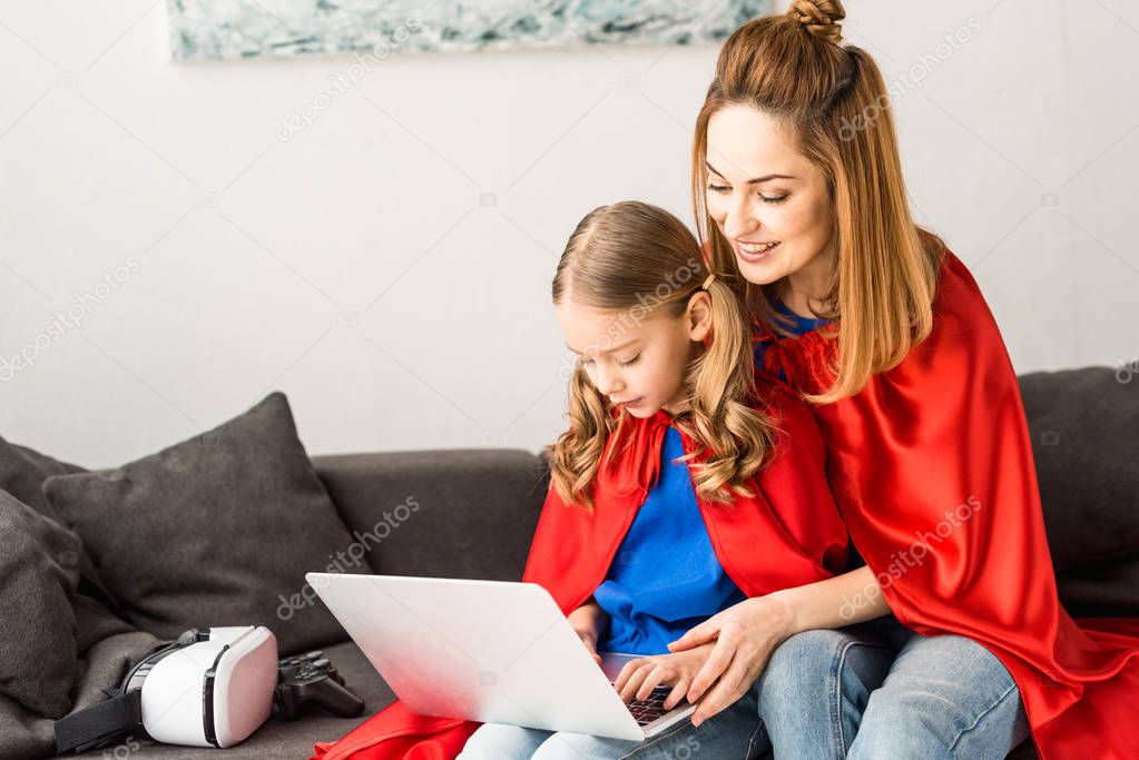 mother and daughter in red cloaks sitting on sofa and typing on laptop